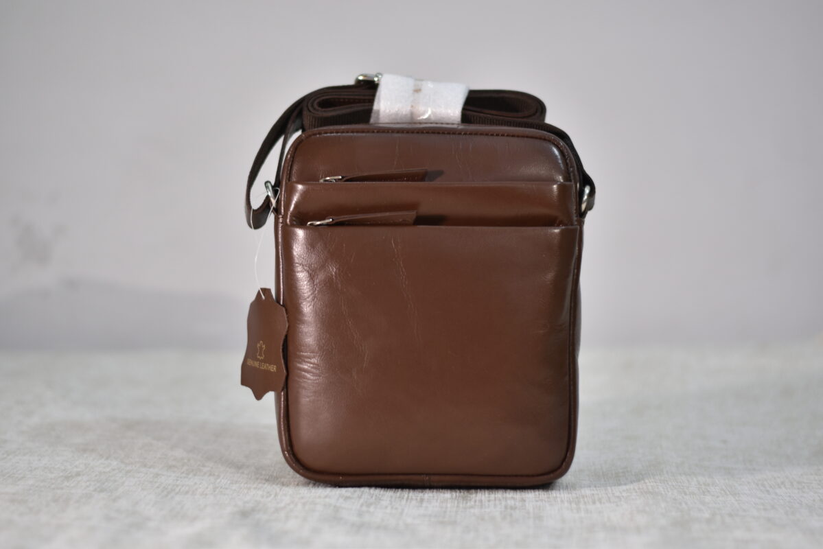 A close-up, front-facing shot of a brown leather crossbody bag with a shoulder strap and multiple zippered compartments, displayed against a plain white background. A small leather tag is visible on the bottom left of the bag.