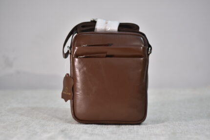 A close-up, front-facing shot of a brown leather crossbody bag with a shoulder strap and multiple zippered compartments, displayed against a plain white background. A small leather tag is visible on the bottom left of the bag.